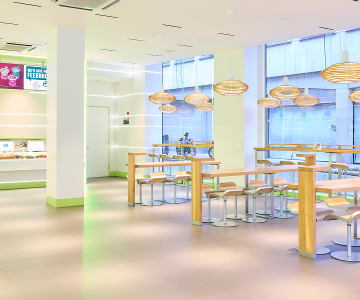 Bright, modern cafe with wooden tables and stools, illuminated by unique pendant lights. Large windows allow natural light to fill the space. A counter with food items is visible in the corner near a colorful sign.