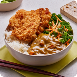 A bowl of white rice topped with two pieces of golden-brown fried chicken, curry with vegetables, and a side of sautéed greens with carrots. Chopsticks are placed beside the bowl on a light green napkin.