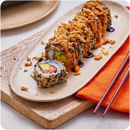 A plate of sushi rolls topped with crispy flakes and drizzled with sauces, placed on a wooden board with a folded orange napkin and chopsticks beside it.