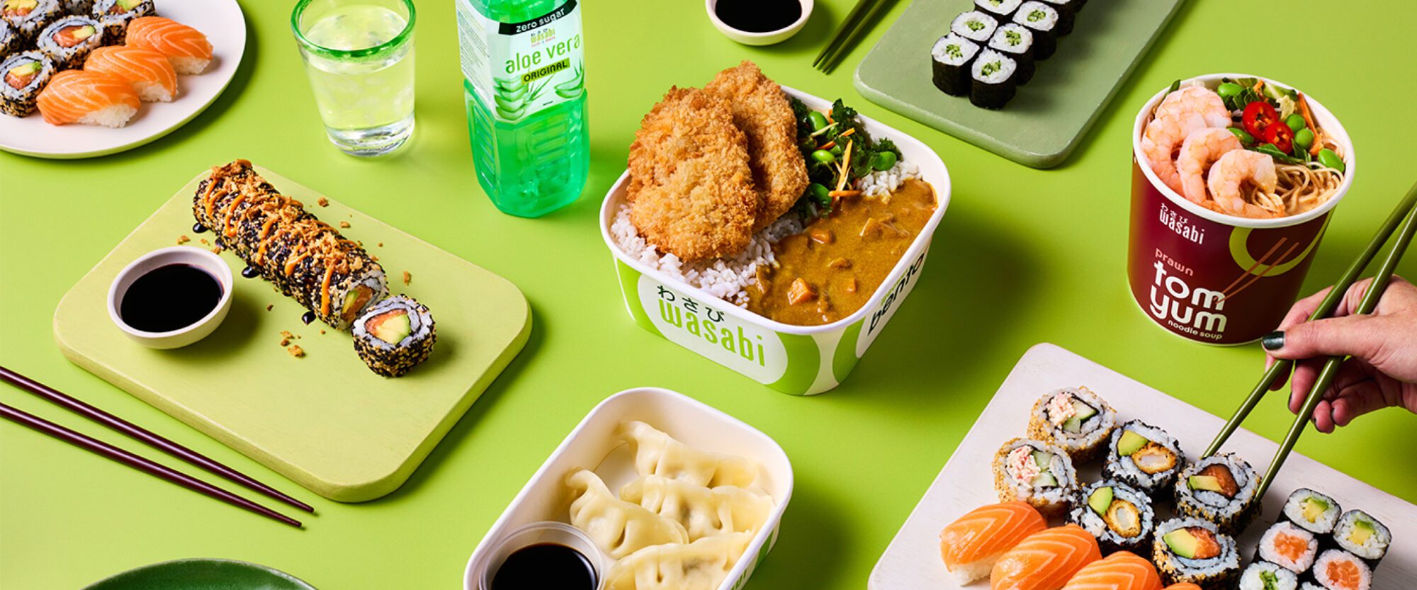 A colorful spread of Asian cuisine on a green background, featuring sushi rolls, salmon sashimi, dumplings, and a bowl of katsu curry with rice. Also shown are a drink bottle, a glass of lemon-lime soda, and a bowl of tom yum soup.