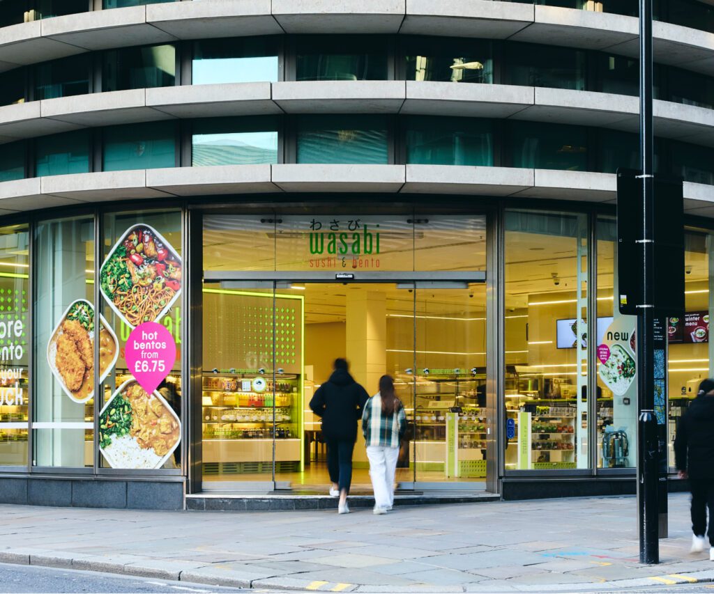 Street view of the entrance to a Wasabi sushi and bento restaurant. Two people enter through glass doors, with vibrant food images displayed on the exterior. A sign highlights a hot bento offer for £6.75.