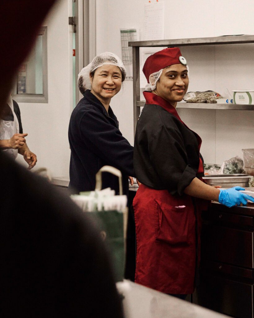 Two people working in a kitchen, wearing hairnets and red uniforms. They are standing beside each other, smiling at the camera. One person is wearing blue gloves and appears to be preparing food. Kitchen equipment and supplies are visible in the background.