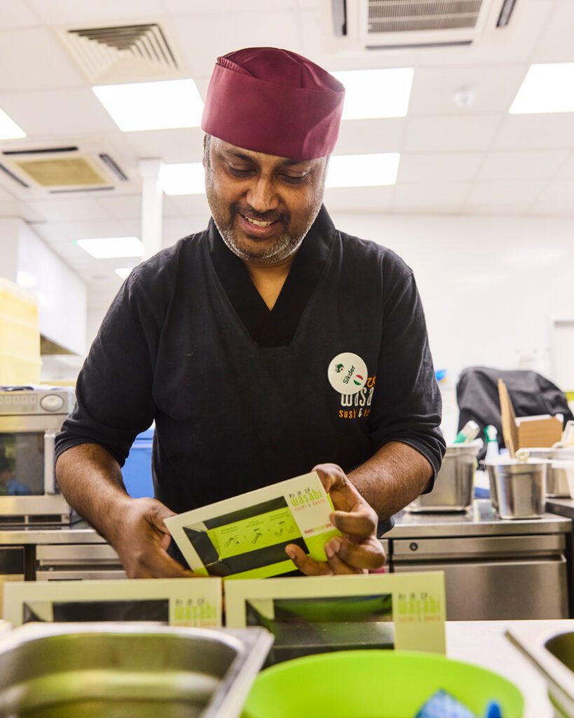 A person wearing a maroon hat and black uniform is smiling while packing sushi boxes in a kitchen. There are various kitchen utensils and appliances in the background.