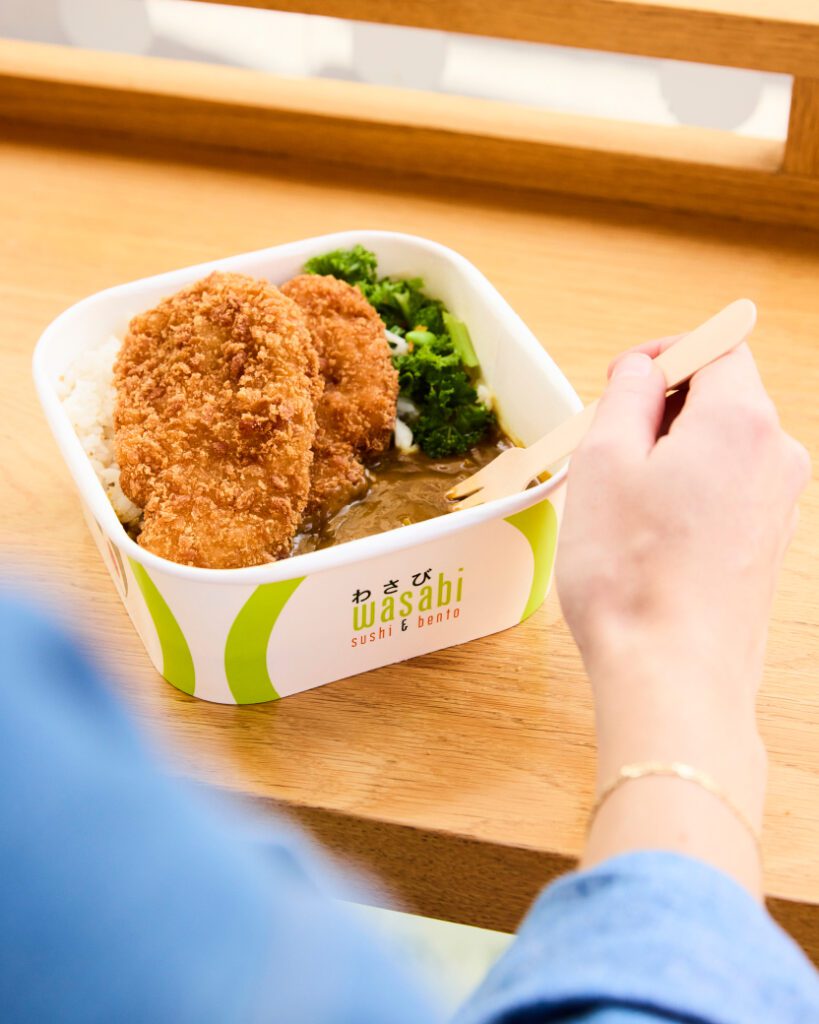 A person holds a wooden fork over a bento box containing rice, breaded chicken cutlets, and broccoli. The box has a 