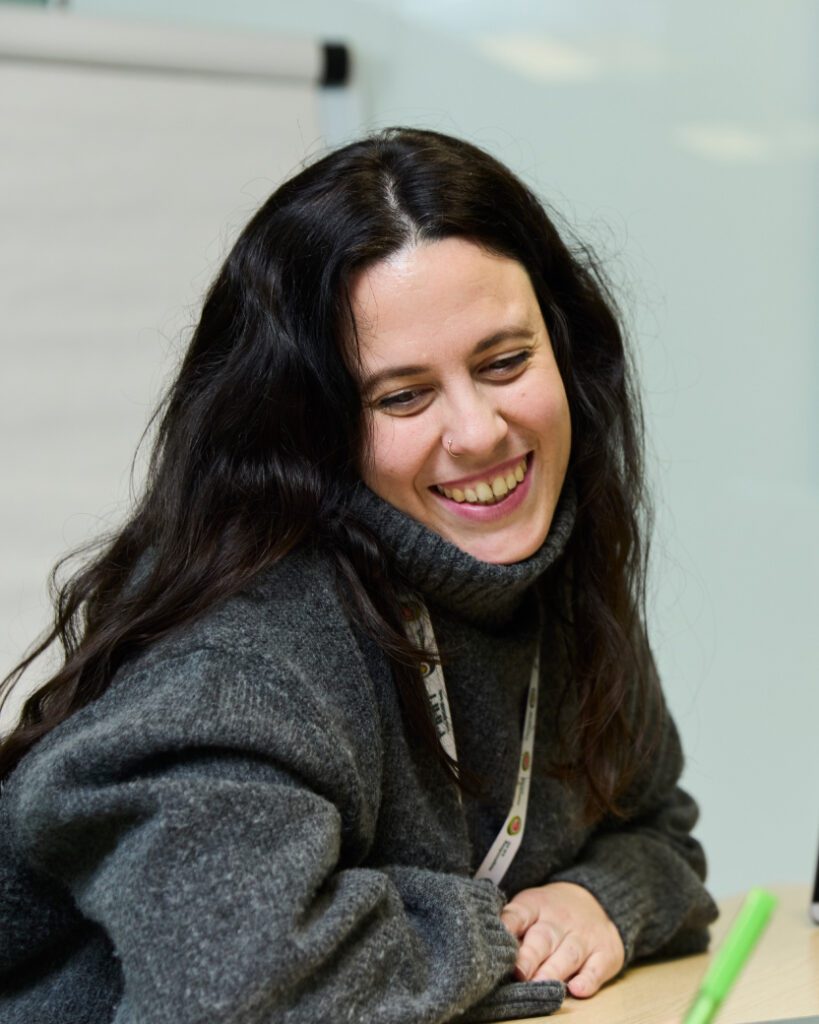 A person with long dark hair, wearing a gray sweater and lanyard, smiling and looking down. The setting appears to be indoors, possibly in a workplace or classroom environment.