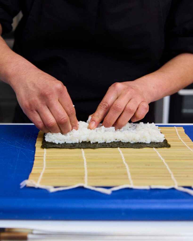 A person preparing sushi by spreading rice on a sheet of nori placed on a bamboo rolling mat. The hands are carefully arranging the rice, with a blue surface beneath the mat.