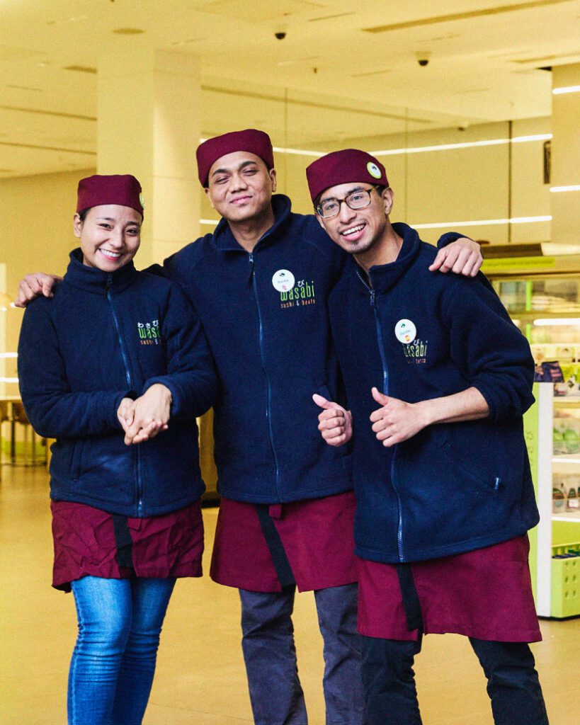 Three smiling restaurant employees wearing matching uniforms and maroon hats pose together inside a brightly lit establishment. They each display friendly gestures, with two giving thumbs up. The atmosphere appears welcoming and casual.