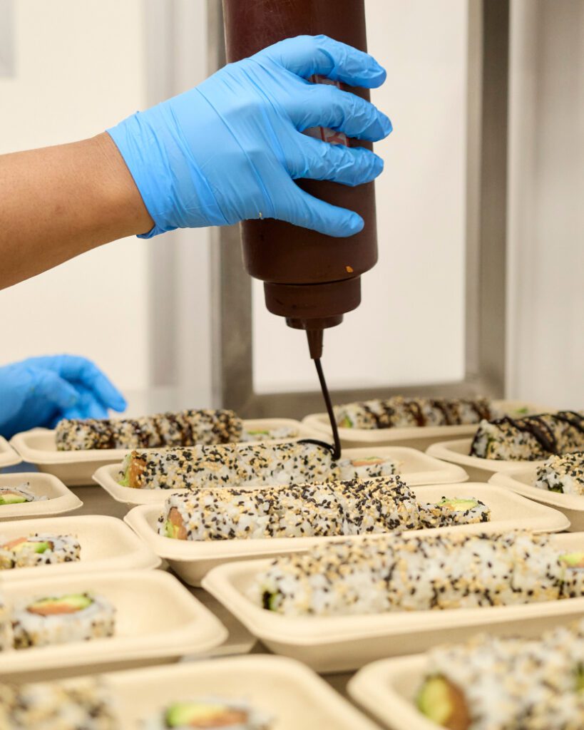 A person wearing blue gloves drizzles sauce over trays of sushi rolls. The sushi is placed in eco-friendly containers and topped with sesame seeds.