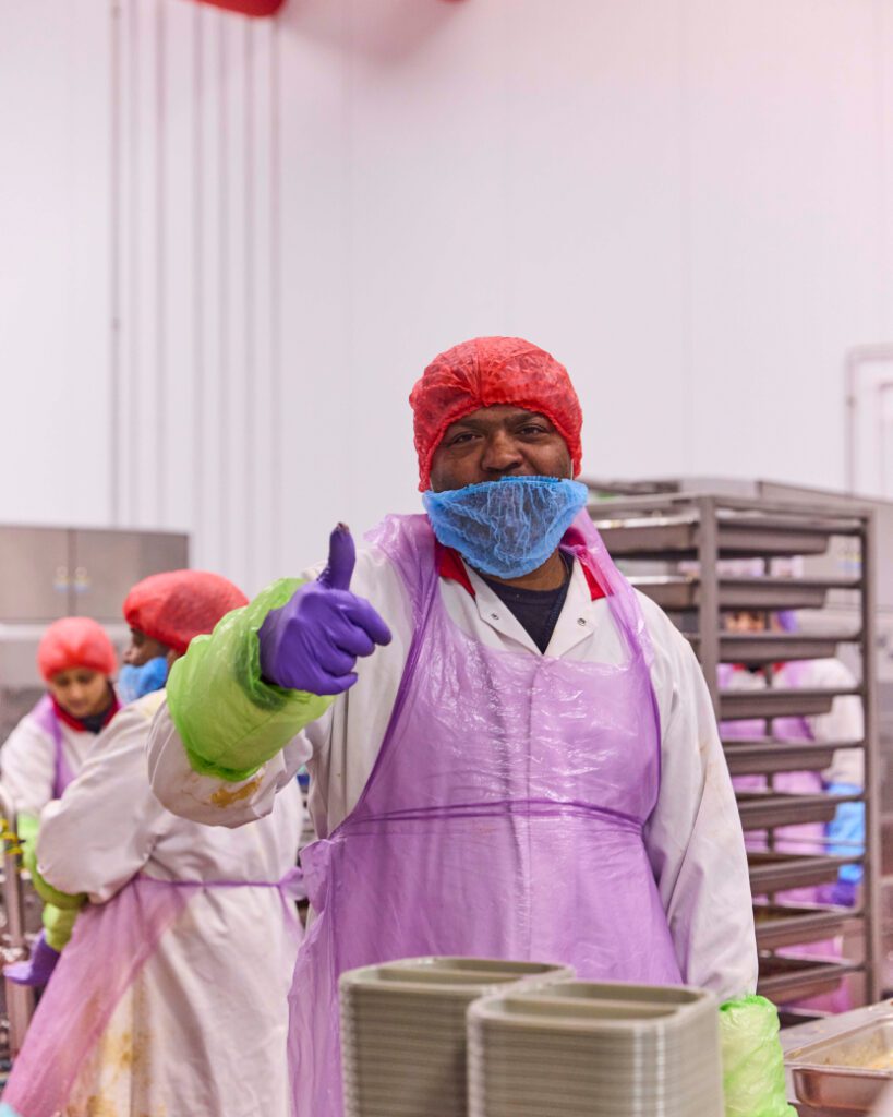 A worker in a food processing facility gives a thumbs-up. The person is wearing a red hairnet, blue beard cover, purple gloves, and a purple apron over a white uniform. In the background, other workers are visible along with metal trays.