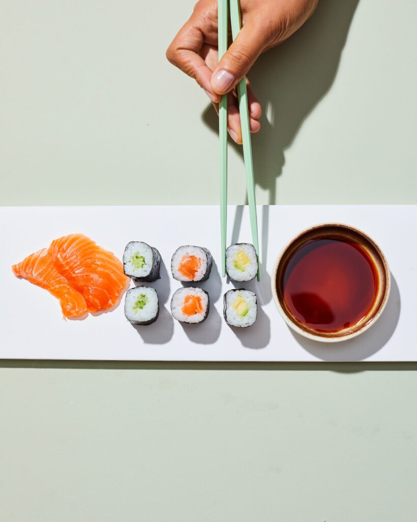 A hand uses green chopsticks to pick up a sushi roll from a white rectangular plate. The plate also holds three slices of salmon sashimi and mixed sushi rolls with green and orange fillings, next to a small bowl of soy sauce.