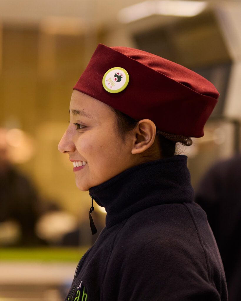 A person wearing a red cap with a smiley face button is shown in profile view, smiling. They are inside a building, with a blurred background, and are dressed in a dark jacket.