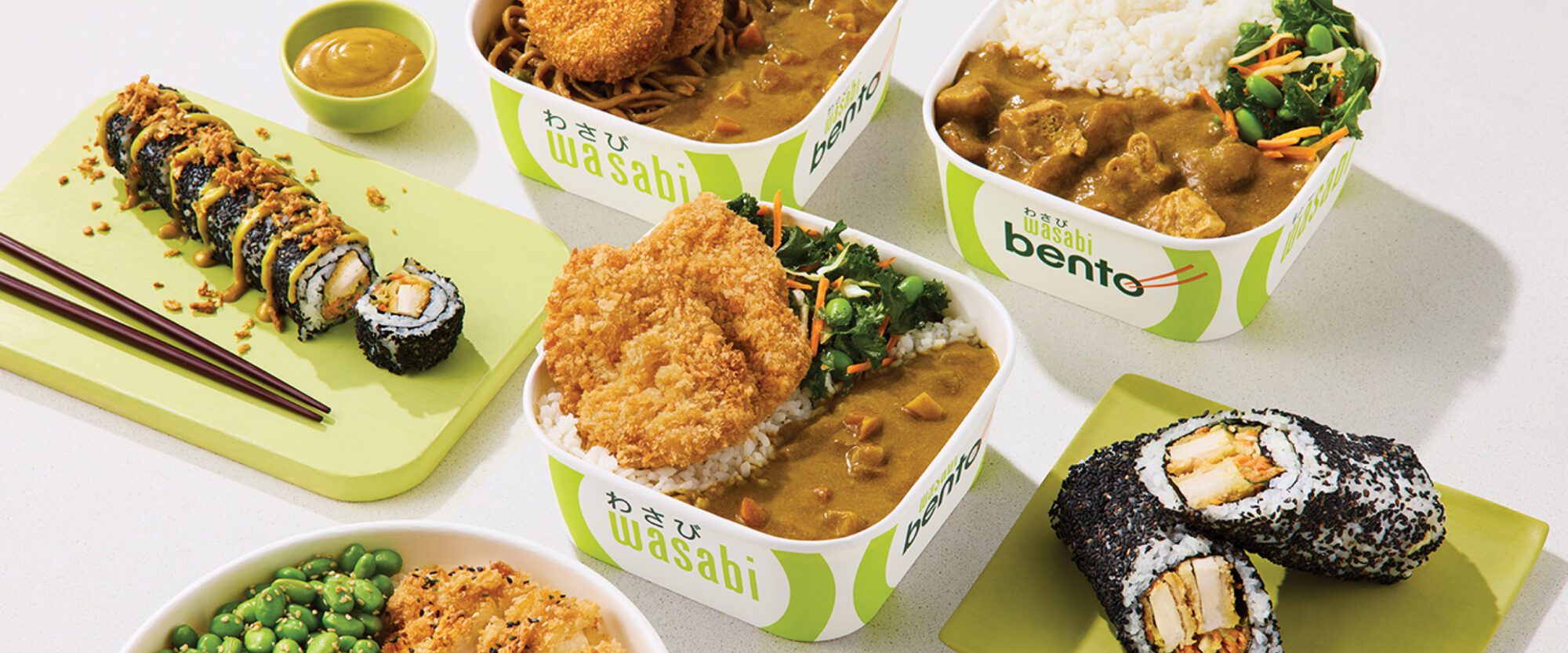 An assortment of Japanese dishes featuring sushi rolls, curry rice with fried cutlets, and soba noodles. Accompanied by edamame and condiments, all served in Wasabi-branded containers on a light surface.