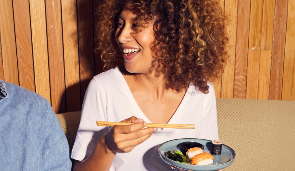 A woman with curly hair smiles while holding a plate of sushi and chopsticks. She's wearing a white shirt and sitting on a couch with a wooden background.