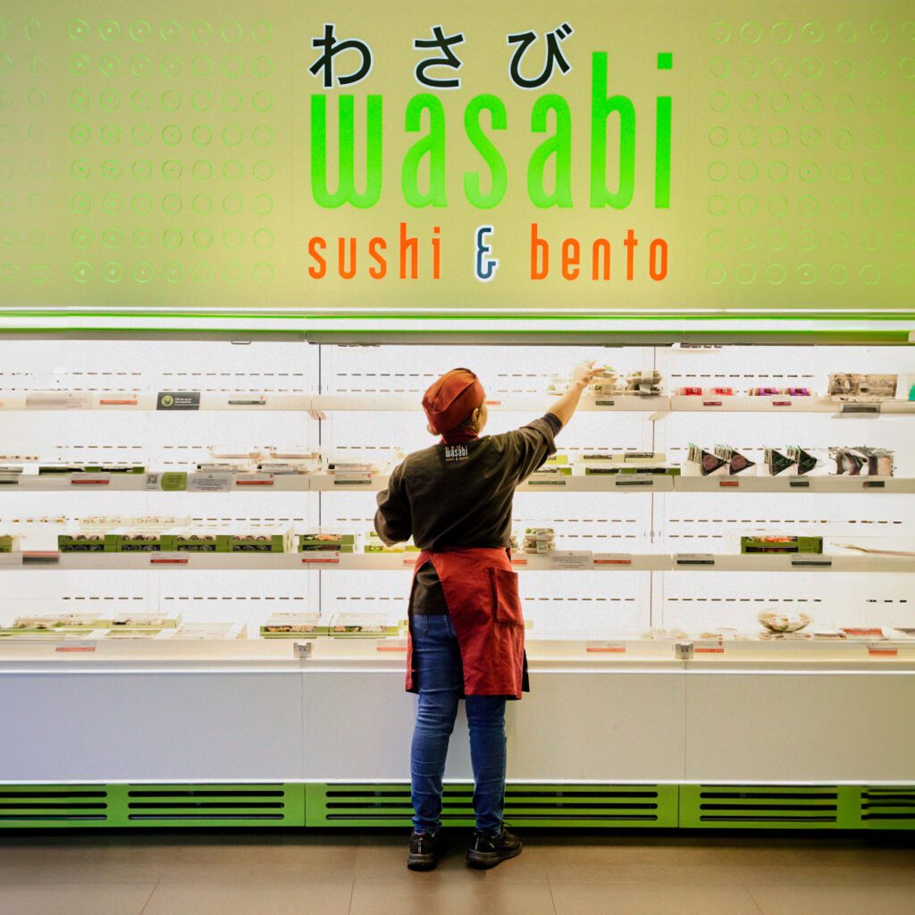 A person in a red apron and hat is arranging items in a well-lit display case at a Wasabi sushi and bento shop. The wall behind them features the Wasabi logo in Japanese and English. The shelves contain various packaged food items.