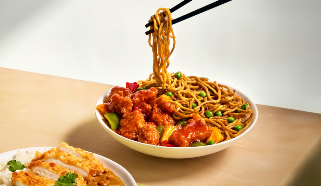 A bowl of noodles with vegetables and saucy fried chicken is being picked up with chopsticks. Another partially visible plate contains fried pieces with sauce on rice, garnished with herbs. Both dishes are on a light wooden surface.