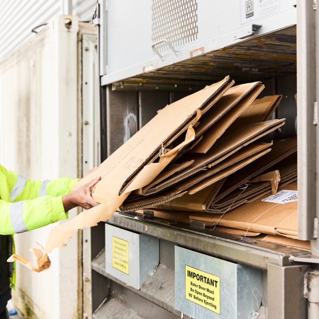 Person in a fluorescent jacket loading flattened cardboard boxes into a recycling compactor outside a building. The compactor has labels, including an important notice about box sizes before closing.