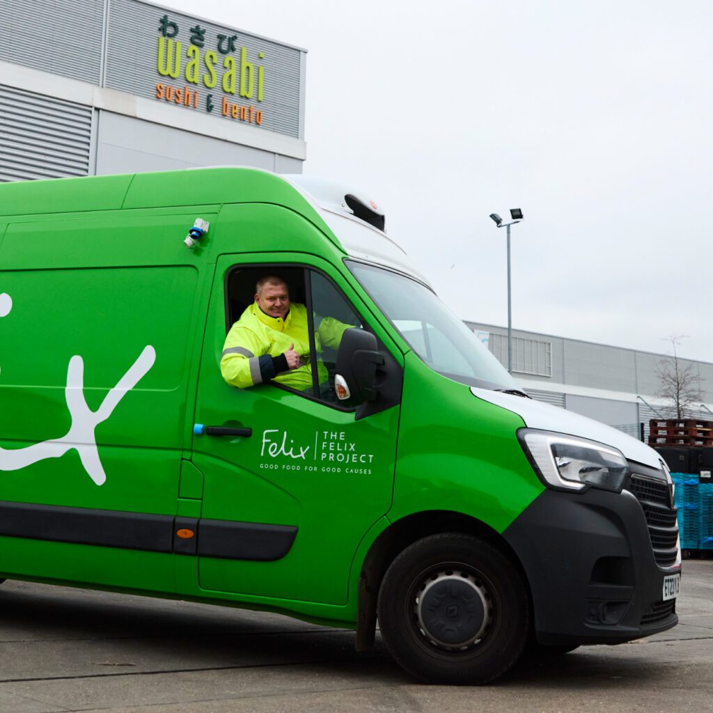 A person in a fluorescent jacket is smiling and giving a thumbs-up from the driver's seat of a bright green Felix Project van, parked outside a building with 