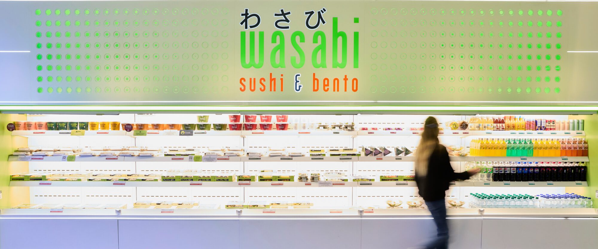 A person walks past a brightly lit display of pre-packaged sushi boxes and drinks at a Wasabi sushi and bento store. The shelves are neatly organized, and a sign with the store's name is visible above.