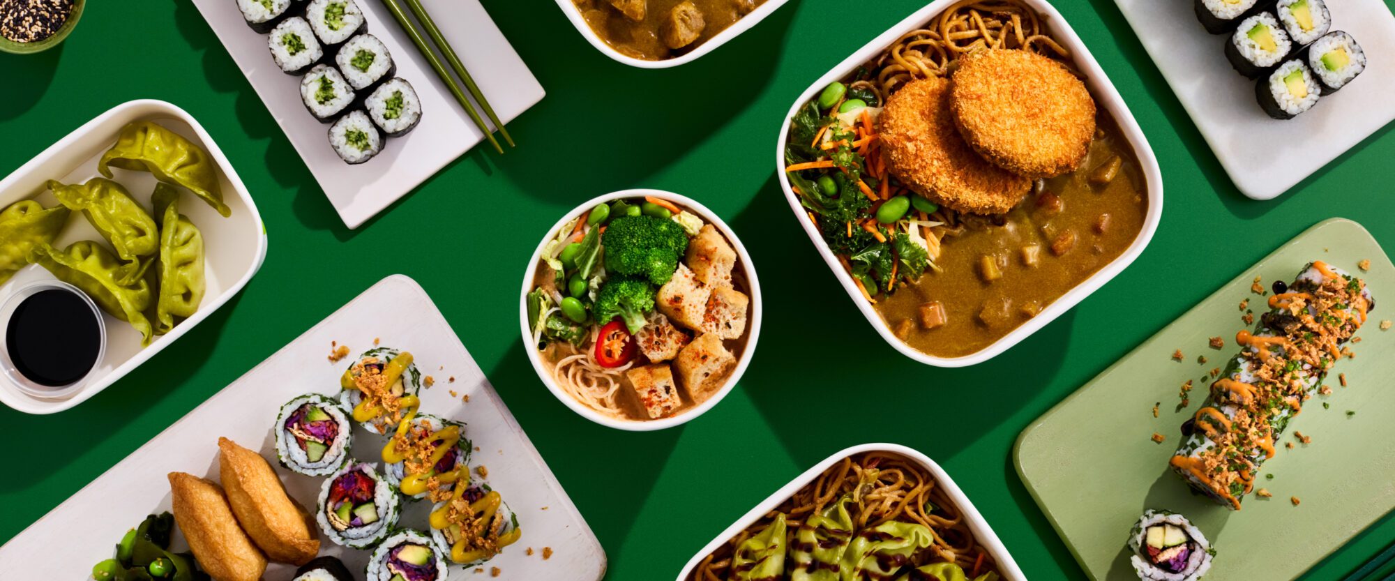 A colorful spread of Asian-inspired dishes on a green background, featuring sushi rolls, ramen with tofu and vegetables, a curry dish topped with crispy patties, and a side of dumplings with soy sauce.