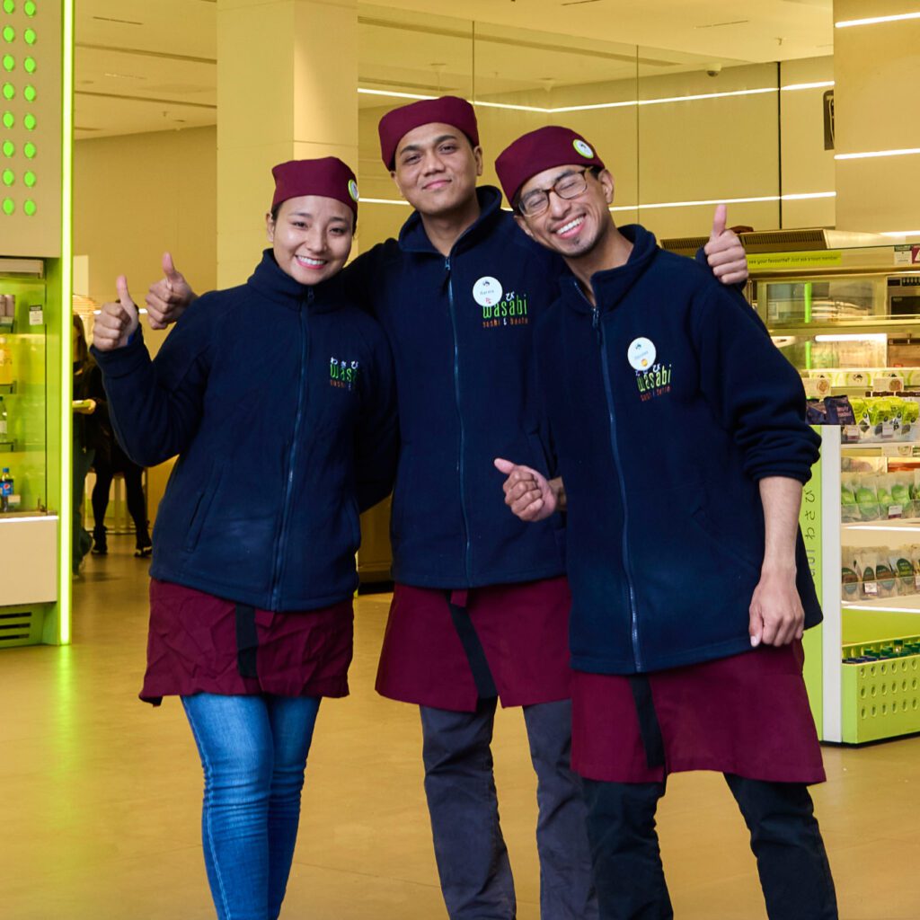 Three people wearing matching uniforms, including red hats and maroon aprons, stand together indoors with thumbs up and smiling. They appear to be in a store or restaurant environment.