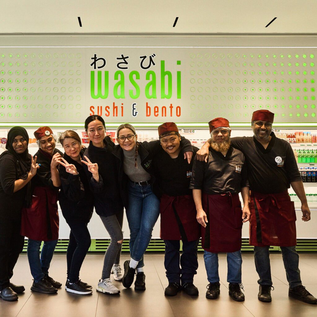 A group of seven people stand smiling in front of a sushi and bento restaurant counter. They are wearing matching uniforms with red aprons and hats. The restaurant sign reads 