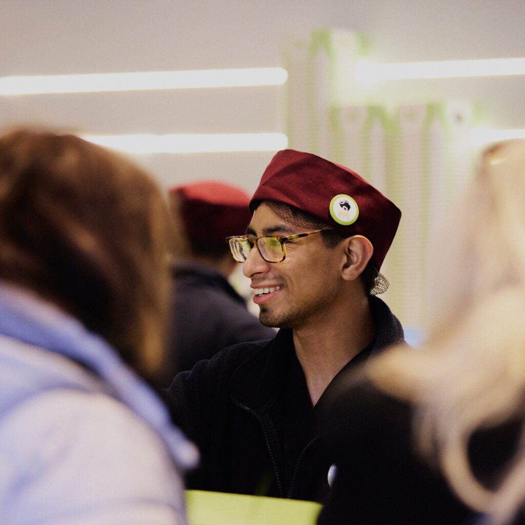 A person wearing glasses and a maroon cap, smiling and talking to someone in a crowded setting. The background has blurred figures and bright lines, suggesting an indoor environment.