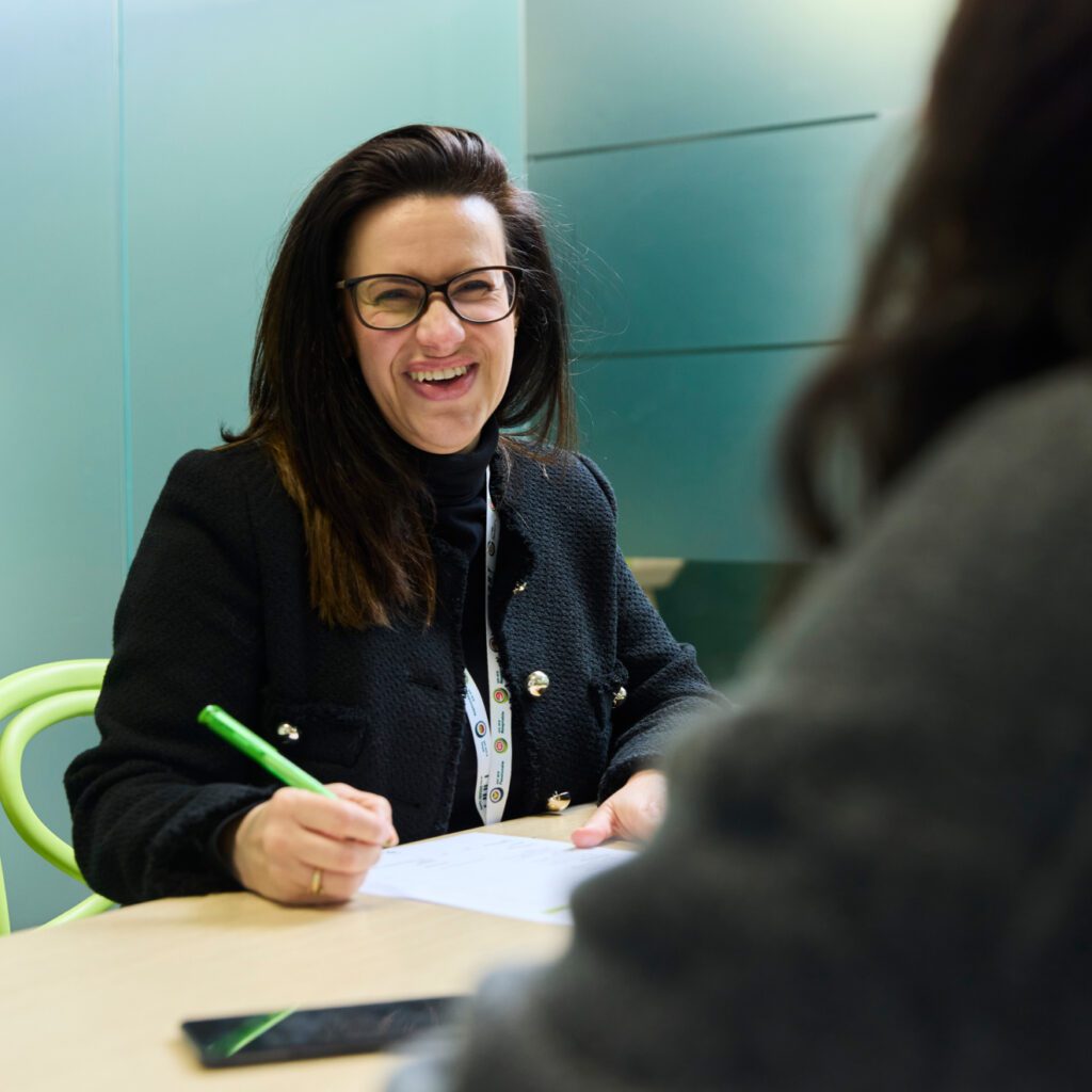 A woman wearing glasses and a black jacket is smiling while holding a green pen. She is sitting at a table, engaged in conversation with another person whose back is to the camera. A smartphone and papers are on the table.