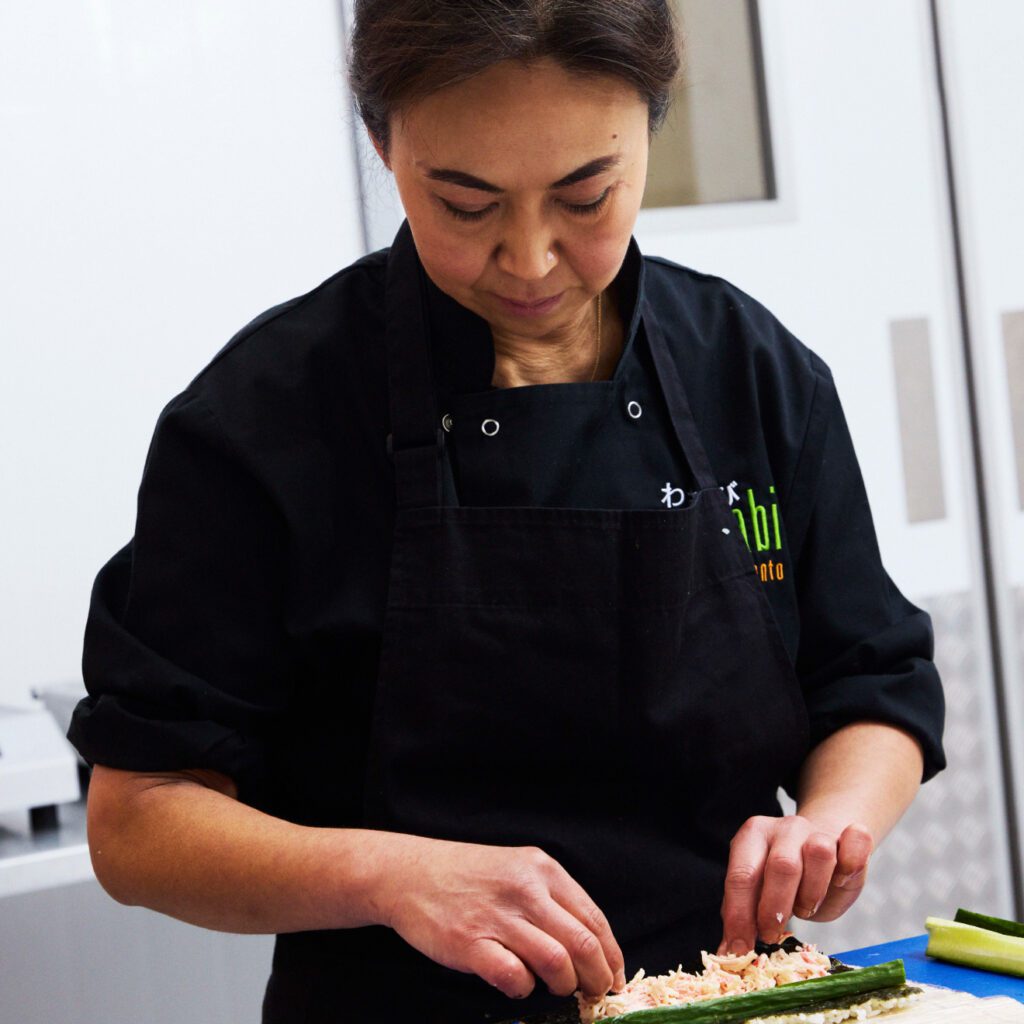 A person wearing a black apron is concentrating on preparing sushi. They are working with ingredients on a blue cutting board in a kitchen setting.