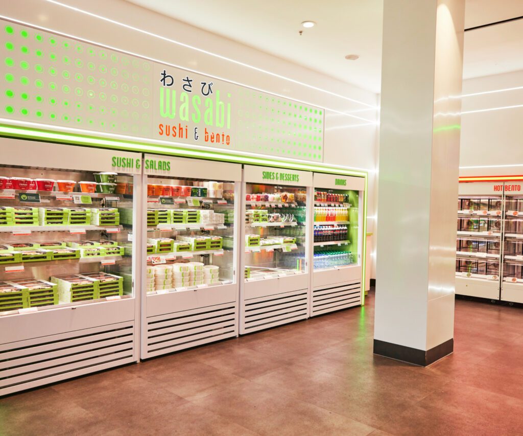 A well-lit store with refrigerated shelves displaying a variety of sushi, salads, desserts, and drinks. The sign above the display reads 