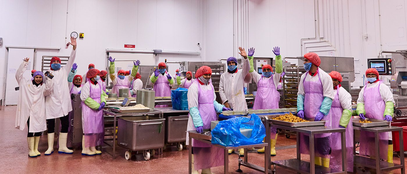 A group of factory workers in colorful protective clothing, including hair nets and gloves, stand in a food processing facility. They cheerfully wave at the camera, surrounded by food trays and equipment.