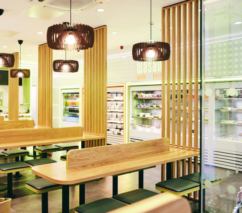 A modern sushi restaurant interior featuring wooden tables and benches. Hanging wooden light fixtures add a decorative touch. Bright green walls and lit refrigerated displays with various sushi and bento boxes line the back of the room.