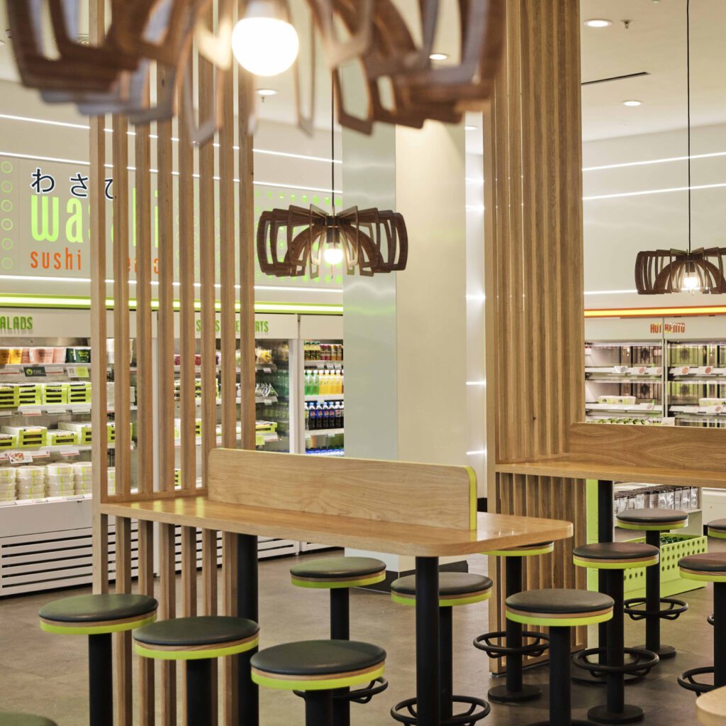 Modern sushi restaurant interior with green and wooden decor. Features high tables with stools and contemporary lighting. Shelves in the background hold various packaged sushi and salads. The ambiance is clean and inviting.