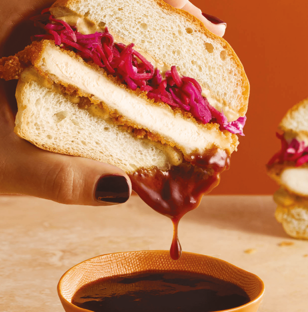 A hand holds a sandwich with fried chicken, vibrant purple coleslaw, and sauce. The sandwich is being dipped into a small bowl of dark sauce, with some dripping down. The background is orange and a similar sandwich is partially visible.