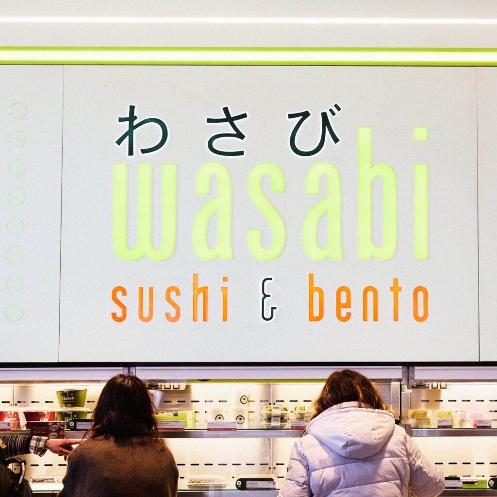 Three people stand in front of a counter at a Wasabi Sushi & Bento restaurant. The sign above them displays the restaurant name in both Japanese and English, with a minimalist design.