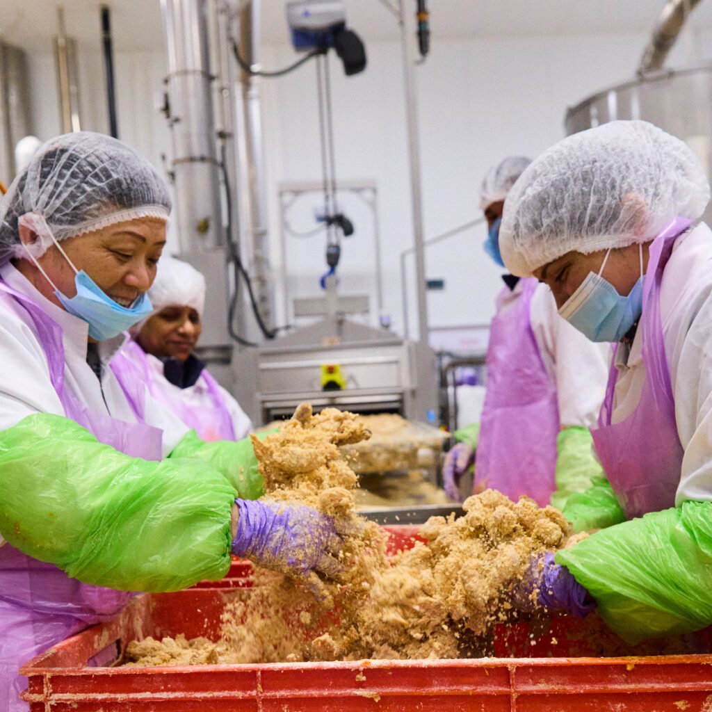 Workers wearing protective gear, including hairnets, masks, gloves, and aprons, mix dough or food ingredients in a large industrial kitchen or food processing facility. The environment is clean and organized, with industrial equipment in the background.