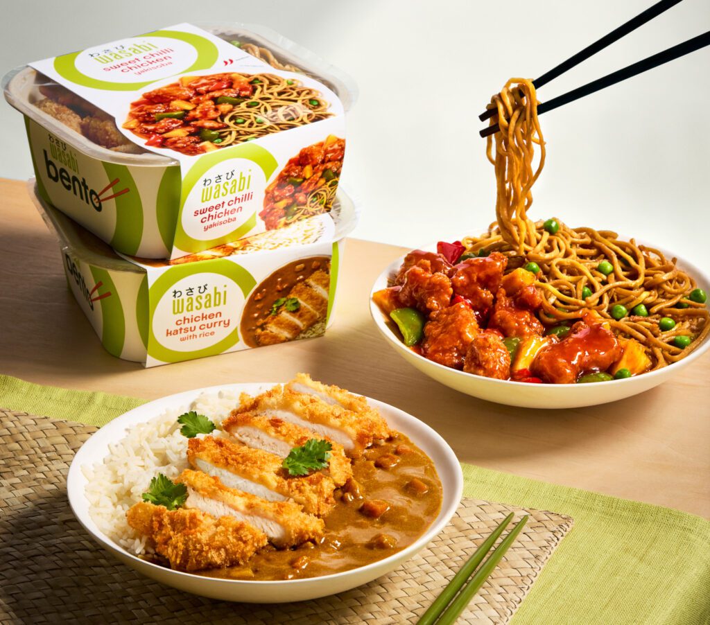 Two bowls of food are shown: one with rice, curry, and breaded chicken; the other with noodles, chicken, and vegetables. Two boxed meals are in the background. A pair of chopsticks lifts noodles from the bowl. A woven mat is partially visible.