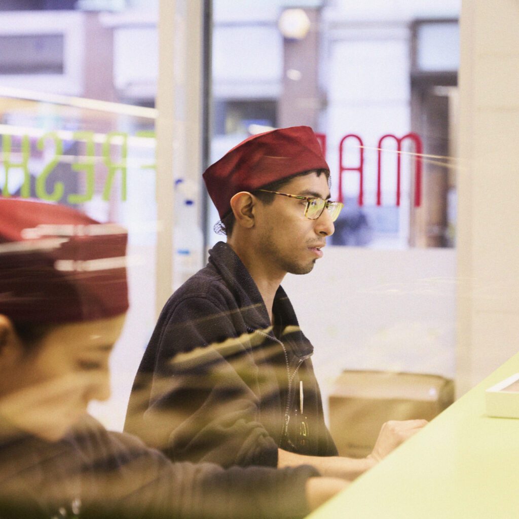 A person wearing glasses and a red cap sits behind a counter, focused on their work. The setting appears to be a shop or restaurant with reflections of the outside visible through the glass.