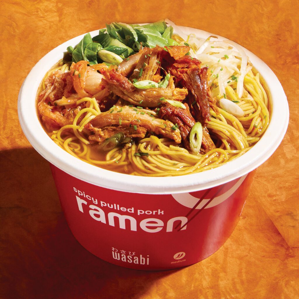 A bowl of spicy pulled pork ramen topped with sliced green onions and leafy greens in a red container labeled 