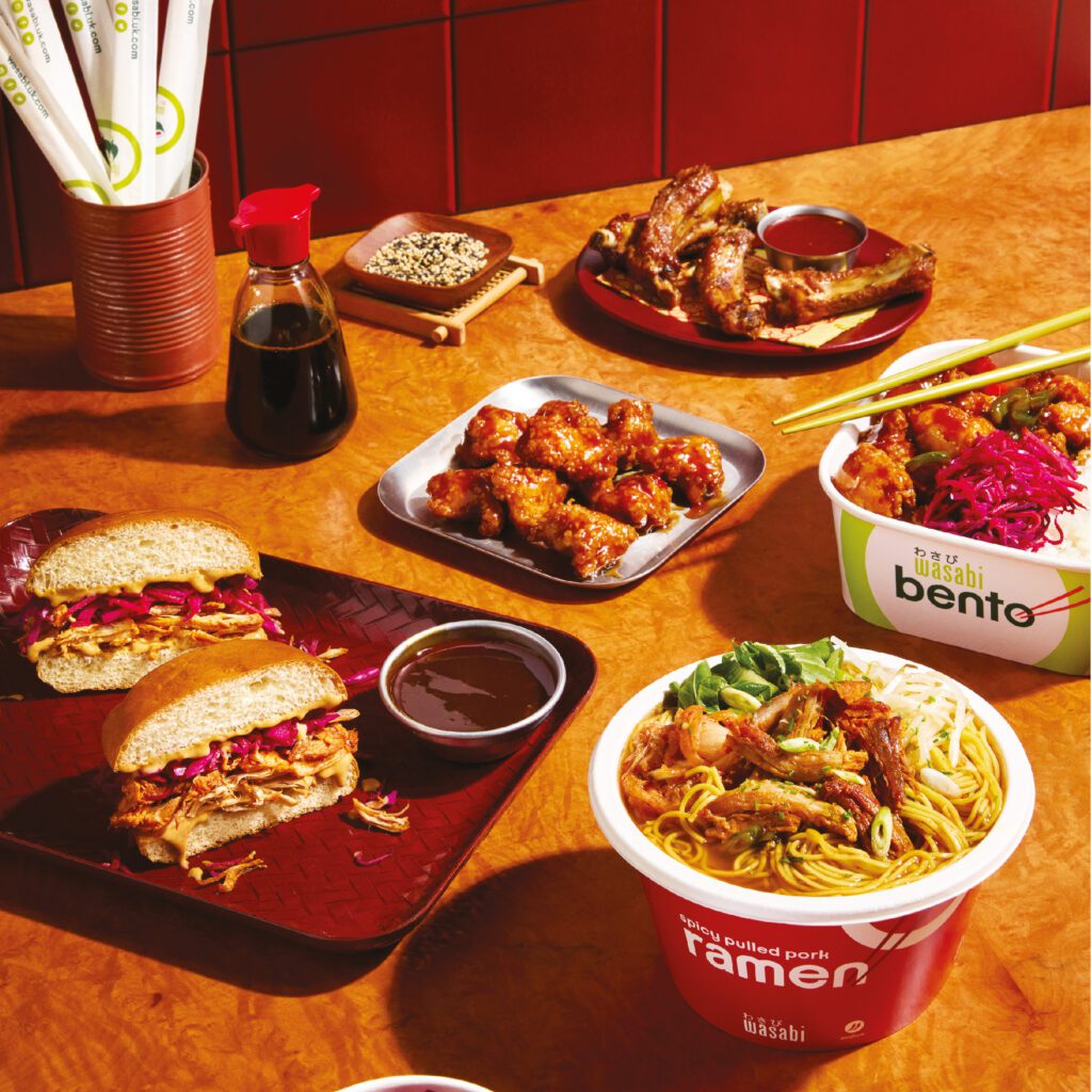 A table with various Asian-inspired dishes, including a bowl of ramen, a pulled pork sandwich with sauce, a plate of glazed chicken wings, and a bento box. Soy sauce, chopsticks, and a spice container are also on the table.