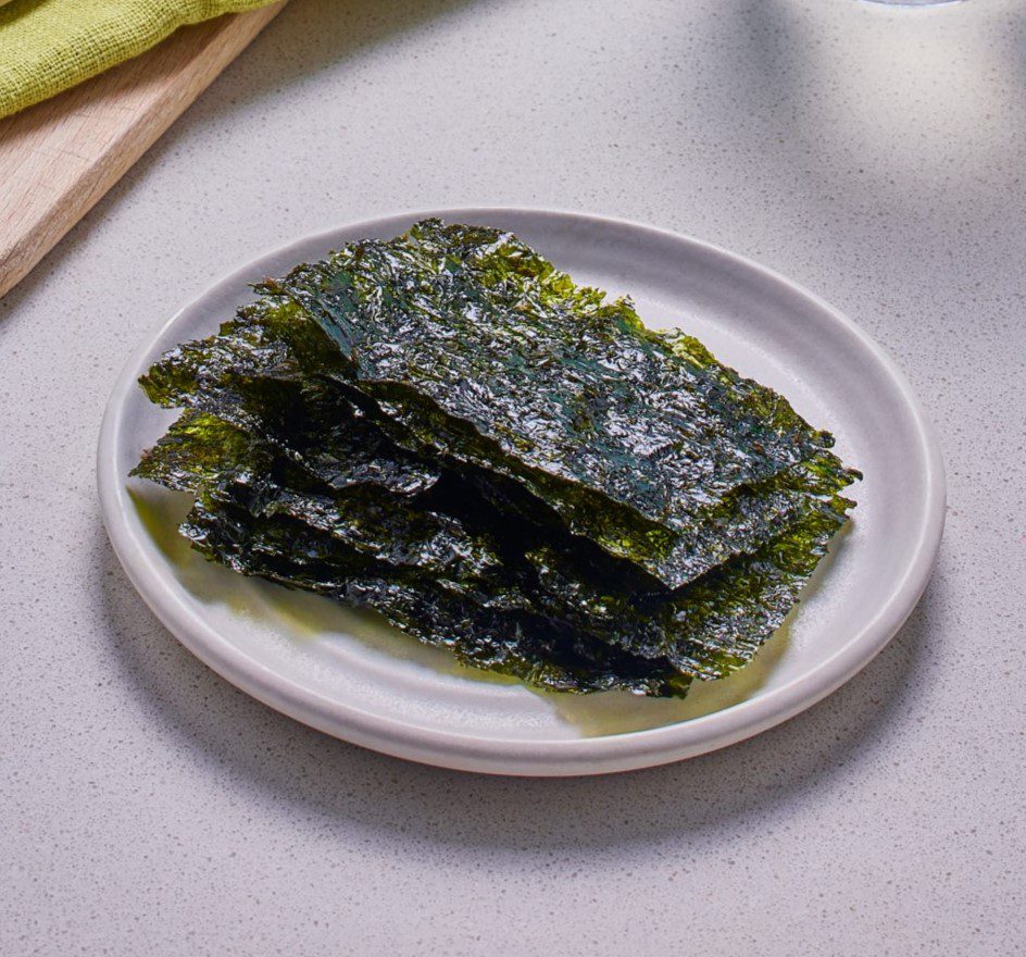 A white plate with a stack of dried seaweed sheets sits on a light-colored countertop. The seaweed has a dark green, glossy appearance. A glimpse of a wooden cutting board and green cloth can be seen in the background.