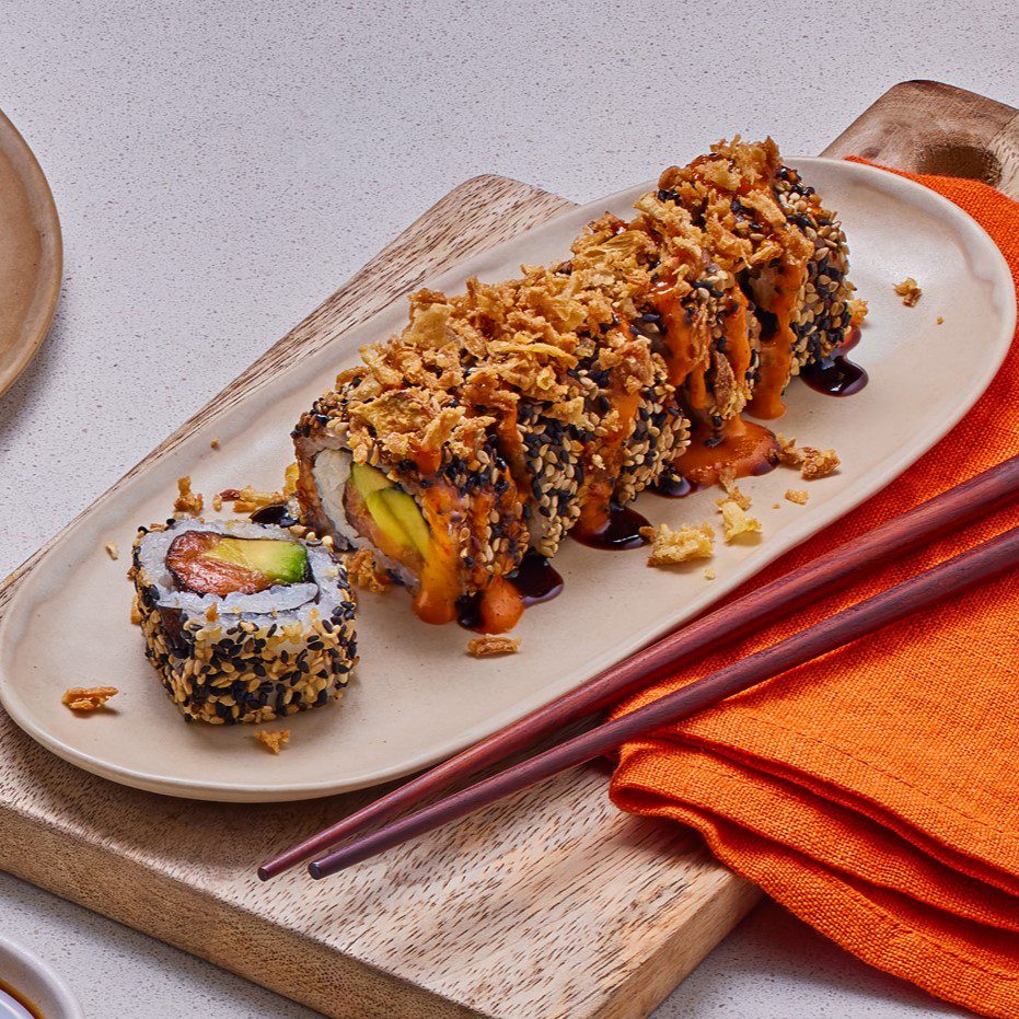 A sushi roll topped with crispy fried onions and drizzled with sauce is placed on a beige oval plate. A piece of sushi is cut and turned to show its colorful filling. Nearby are chopsticks and an orange napkin on a wooden board.